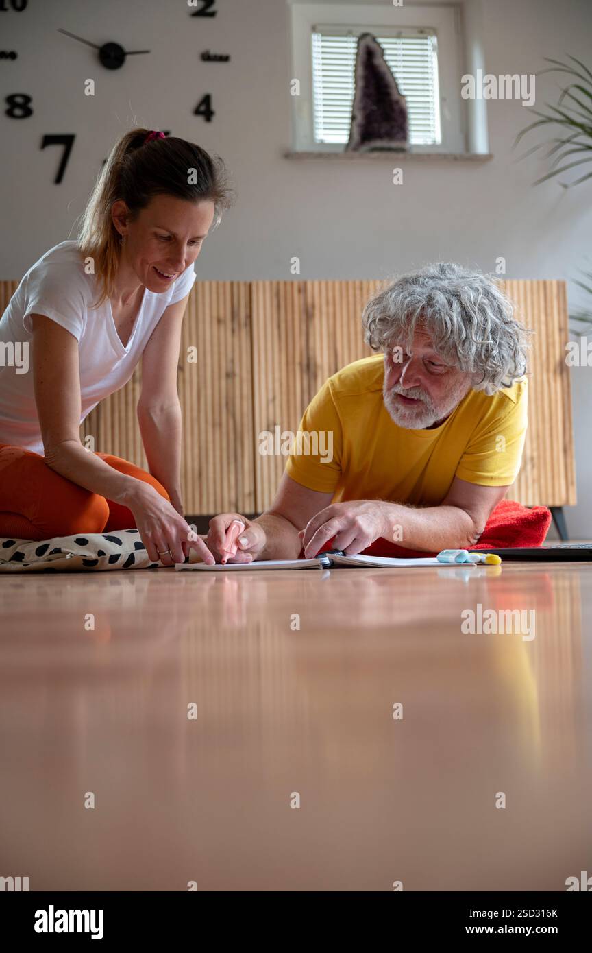 Eine junge Frau und ein älterer Mann arbeiten zusammen an einem Projekt, beide engagiert und lächelnd, während sie auf Papier schreiben, sitzen auf dem Boden in einer gemütlichen, gut beleuchteten Stockfoto