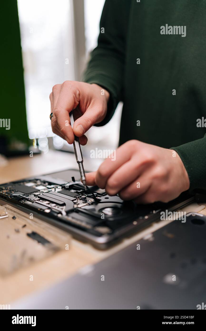 Techniker, männlich, repariert Laptop-Mainboard mit Schraubendreher, umgeben von verschiedenen Komponenten und Werkzeugen auf Werkbank, Nahaufnahme. Computerkonzept Stockfoto