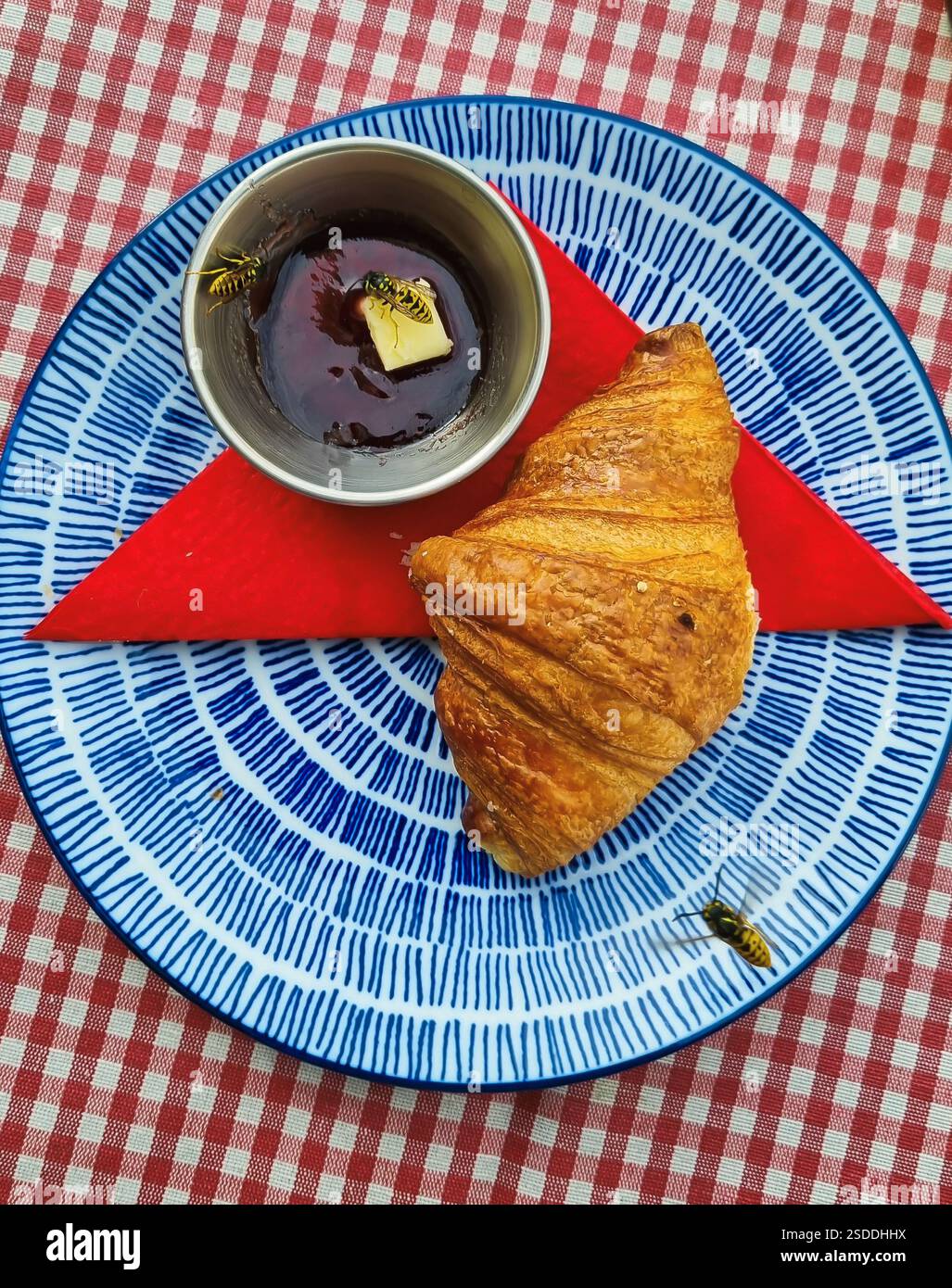 Croissant auf einem bunten Teller auf einer karierten Tischdecke mit einer Tasse Sirup in der Nähe und drei Bienen darüber Stockfoto