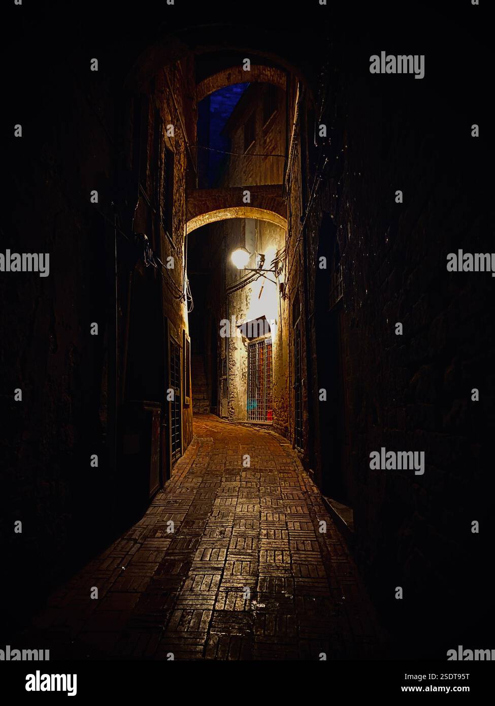 Nachtfoto einer dunklen Straße mit einem beleuchteten Bogen, alten Gebäuden, sanftem Licht und einem Steinboden, der das Licht reflektiert. - Smartphone-aufgenommenes Stockfoto