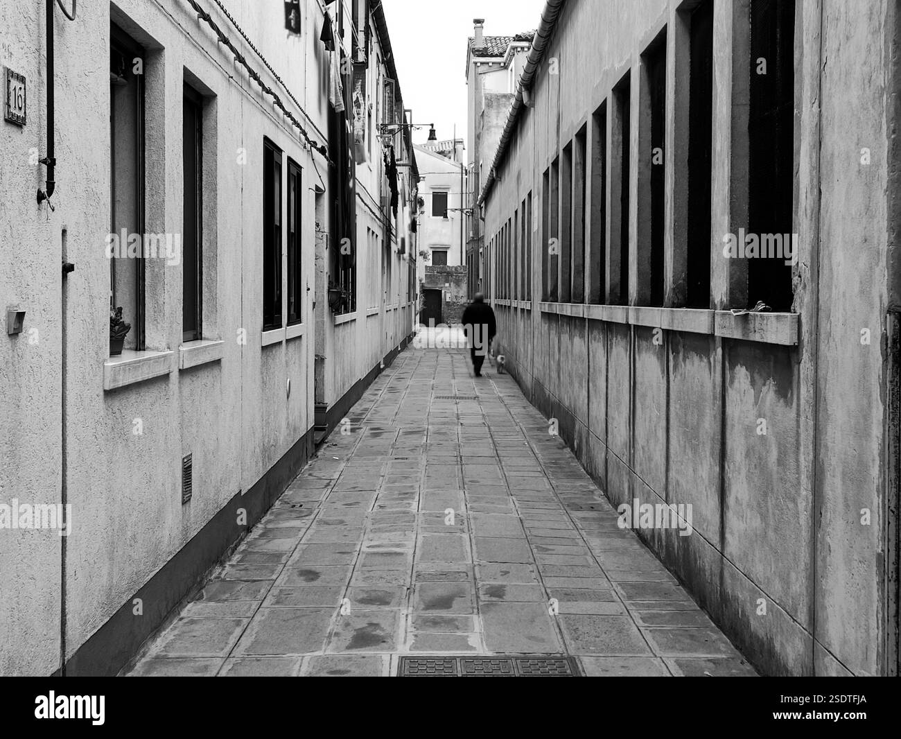Eine Einzelfigur läuft eine enge Gasse entlang, die von alten Gebäuden gesäumt ist. Die Schwarzweißtöne verstärken die Tiefe und die ruhige Atmosphäre Stockfoto