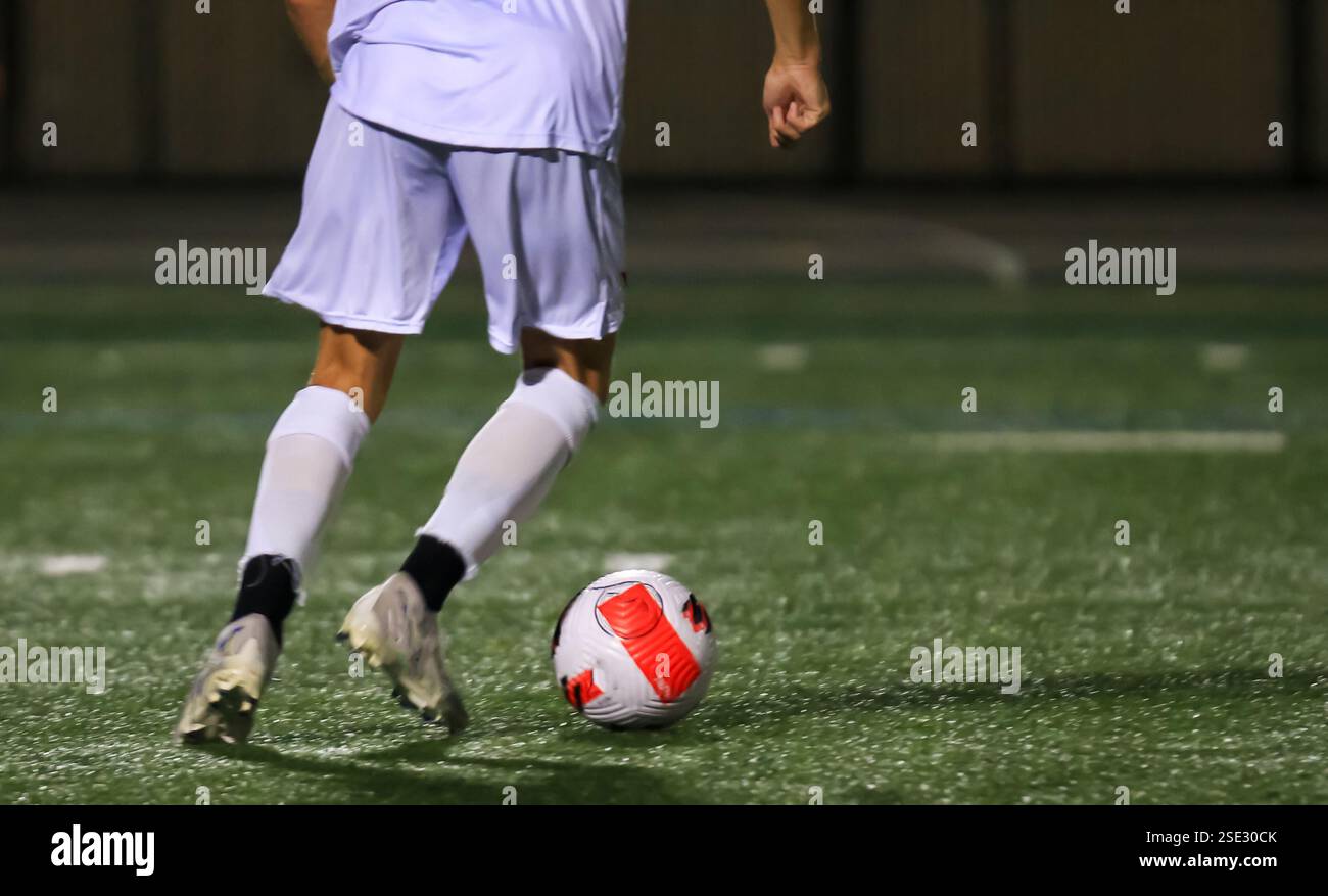 Ein Spieler steuert geschickt einen Fußball auf einem schwach beleuchteten Rasenfeld während eines abendlichen Spiels unter den Lichtern. Stockfoto