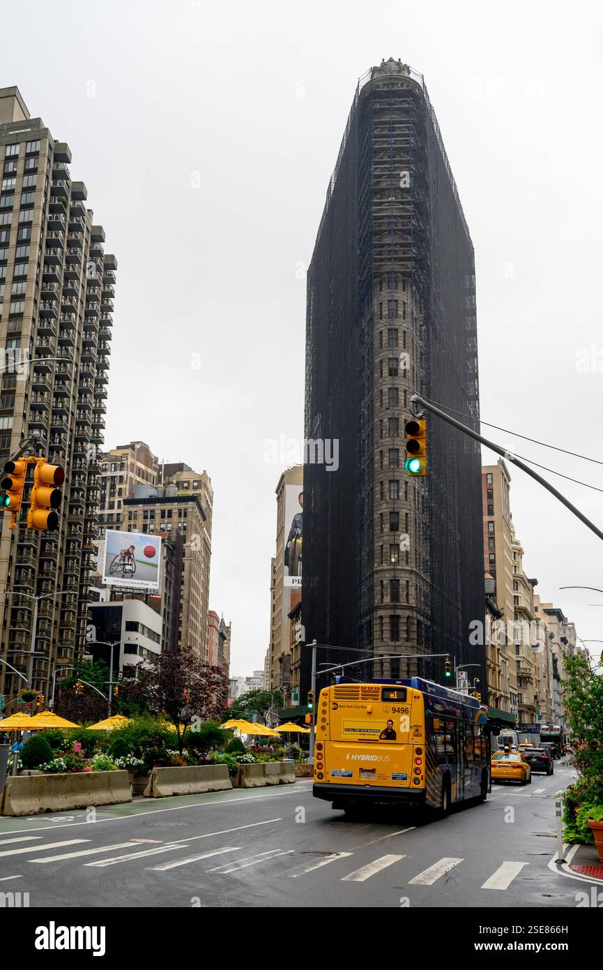 An einem bewölkten Nachmittag zieht das mit schwarzen Gerüsten bedeckte „Flat Iron“-Gebäude die Aufmerksamkeit auf sich, während ein gelber Bus durch die belebte Straße fährt Stockfoto