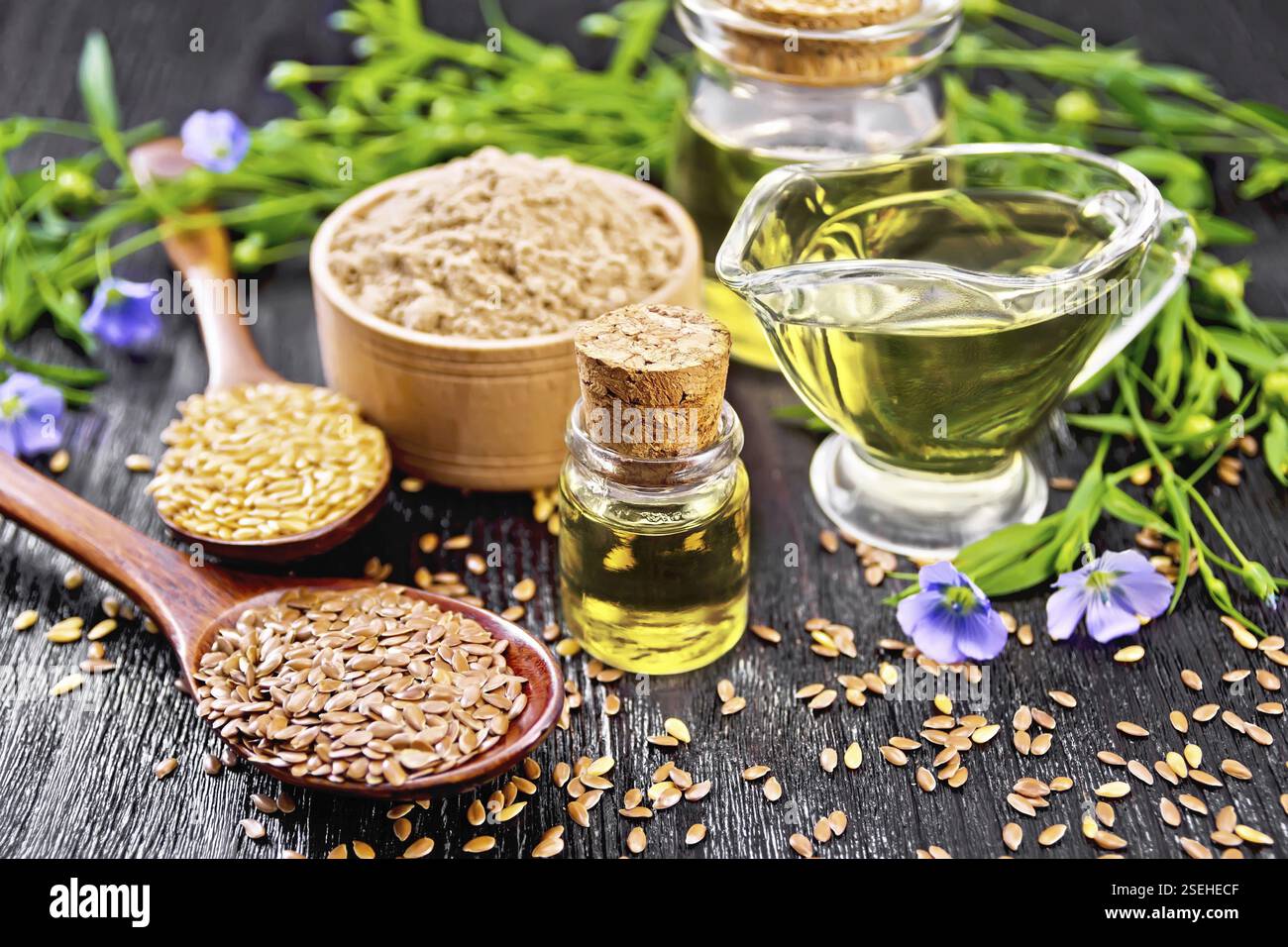 Leinöl in zwei Gläsern und einer Soße Boot mit weißen und braunen Leinsamen in Löffel Mehl in eine Schüssel, Blätter und Blumen auf schwarzem Brett ba Stockfoto