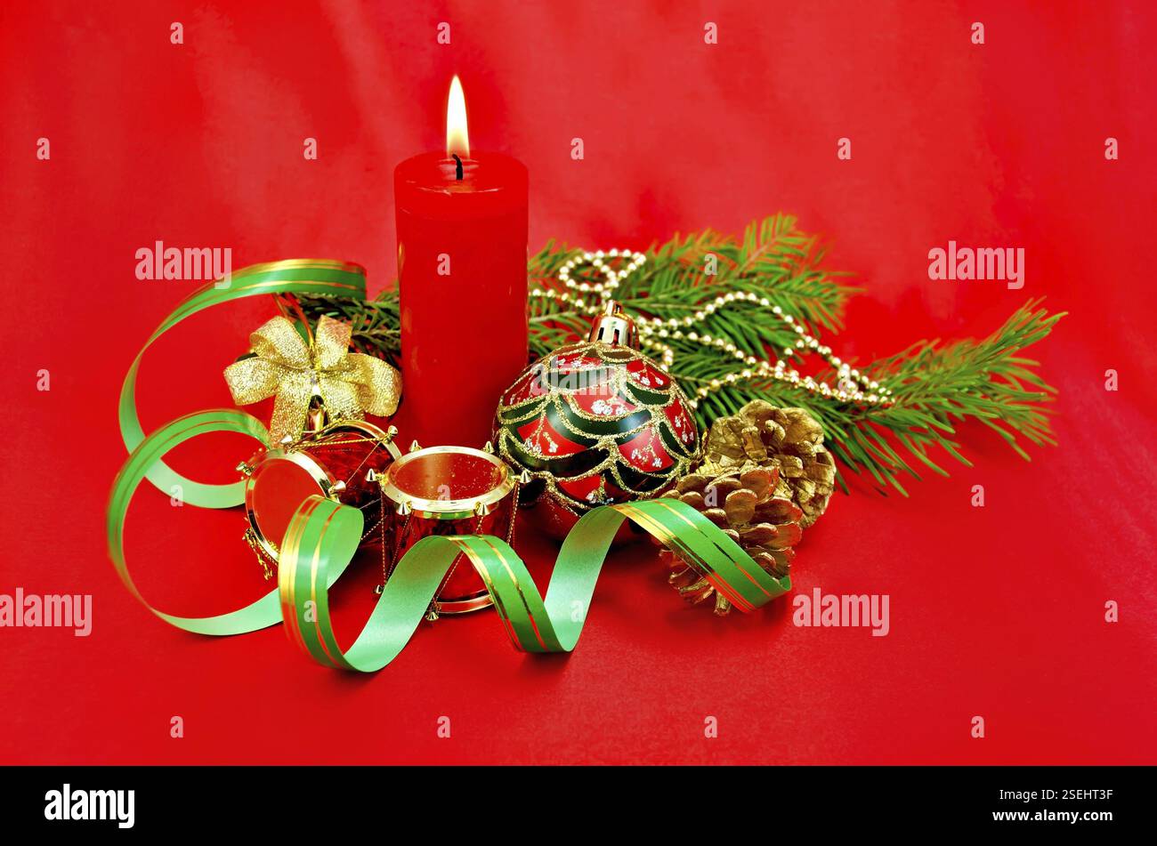 Rote brennende Kerze, Weihnachtsbaumschmuck in Form einer Kugel, eine Handglocke mit Schleife, eine Trommel, Kegel auf roter Seide Stockfoto