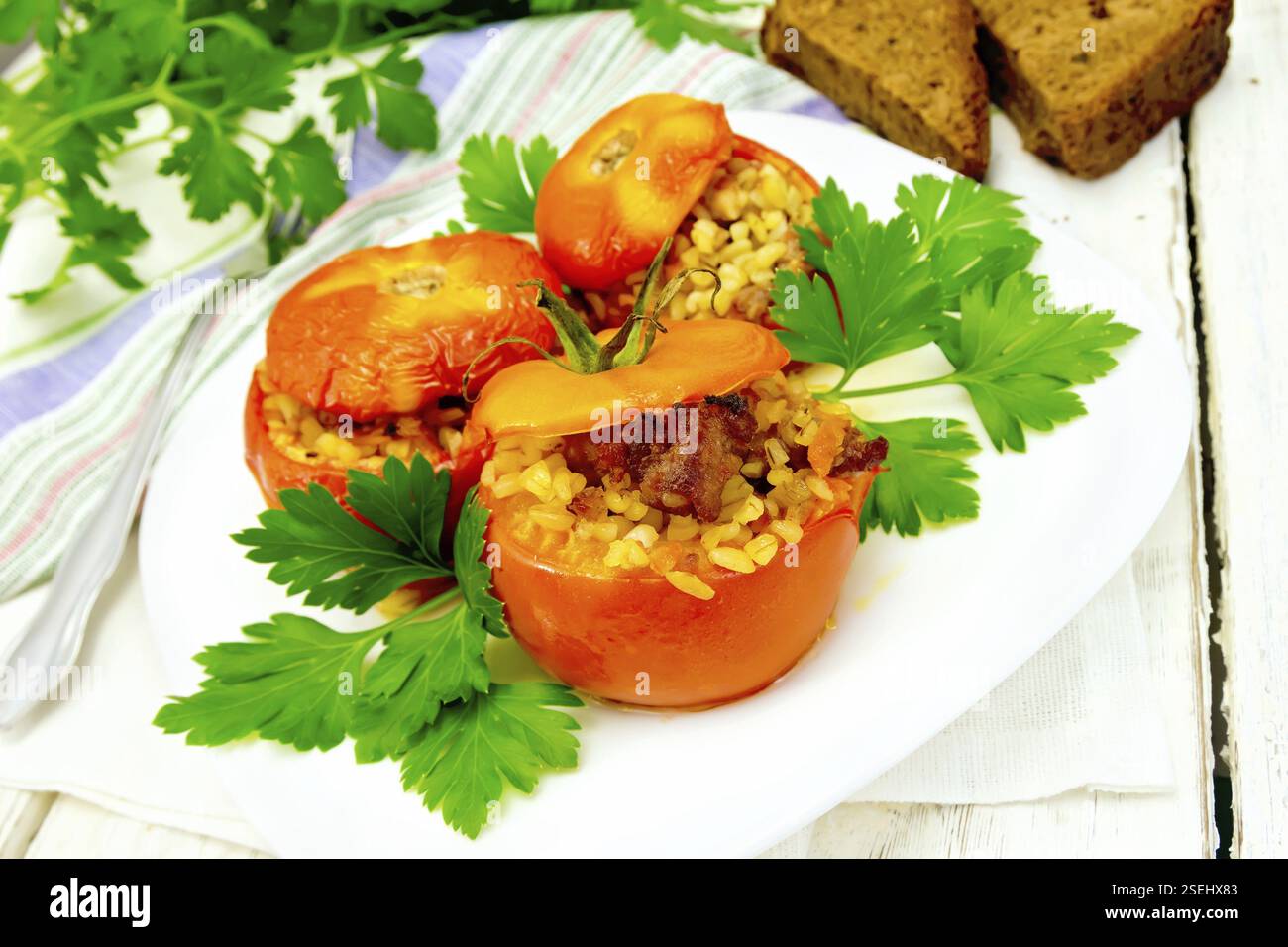 Tomaten gefüllt mit Fleisch und Weizen-Bulgur in einem weißen Teller, Serviette, Gabel, Brot und Petersilie auf den Hintergrund Licht Holzbrettern gedämpft Stockfoto