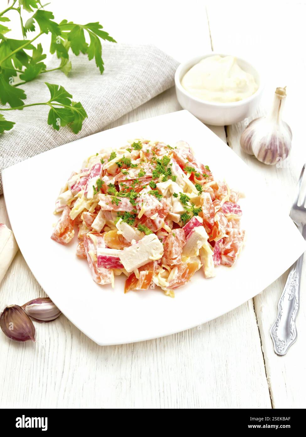 Salat von Crab Sticks, Käse, Knoblauch und Tomaten, mit Mayonnaise, Küchenpapier und Petersilie auf einem Holzbrett Hintergrund Stockfoto