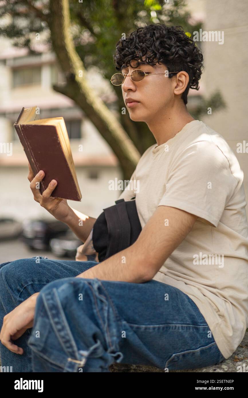Ein Student liest ein Buch außerhalb seiner Bildungseinrichtung und genießt einen ruhigen Moment. Ideal für Themen wie Studium, akademisches Leben und Lesen im Freien Stockfoto