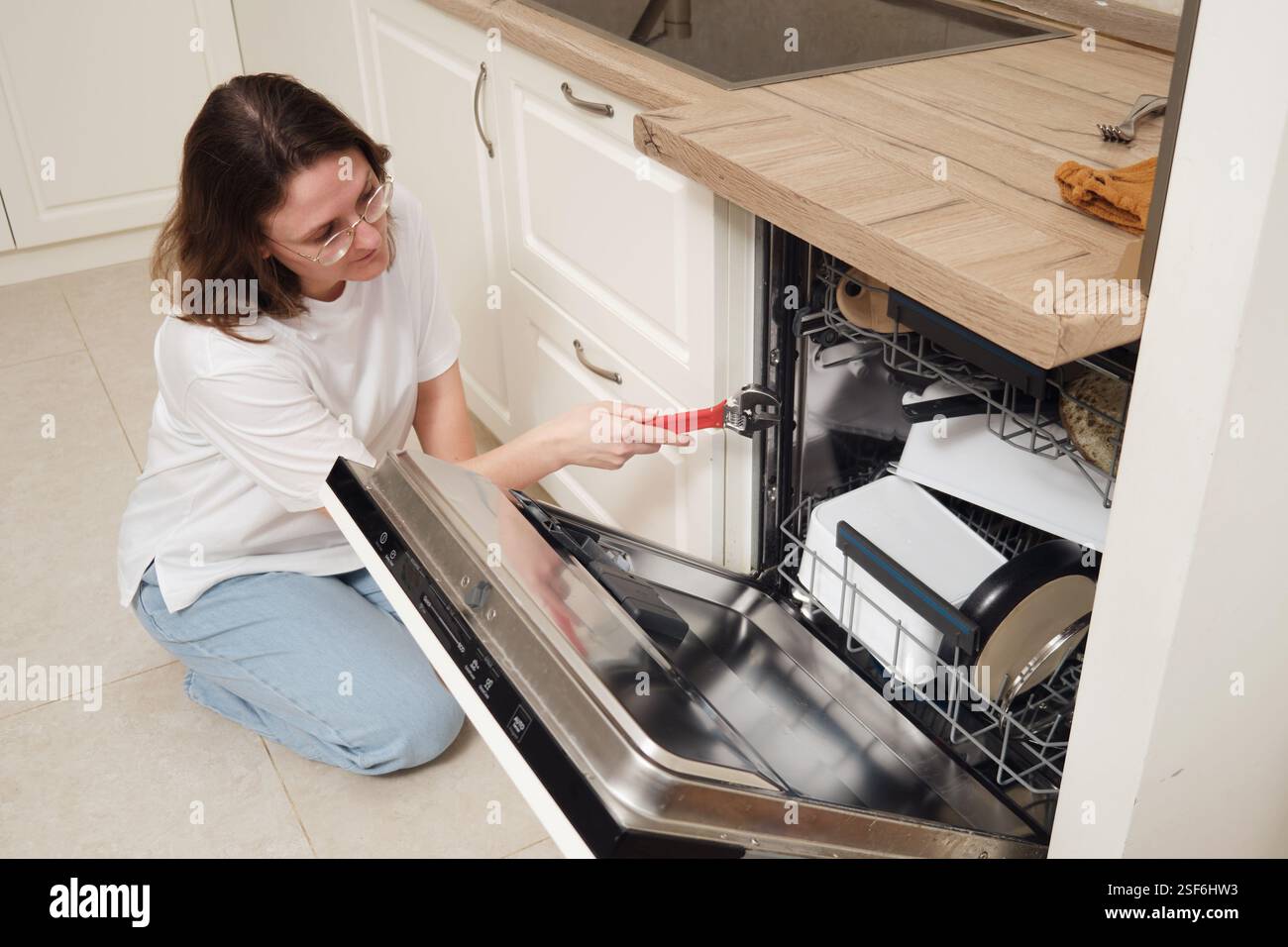 Frau repariert einen Geschirrspüler mit einem Schraubenschlüssel in einer modernen Küche. Haushaltsgerätewartung und Küchenreparaturkonzept. Stockfoto