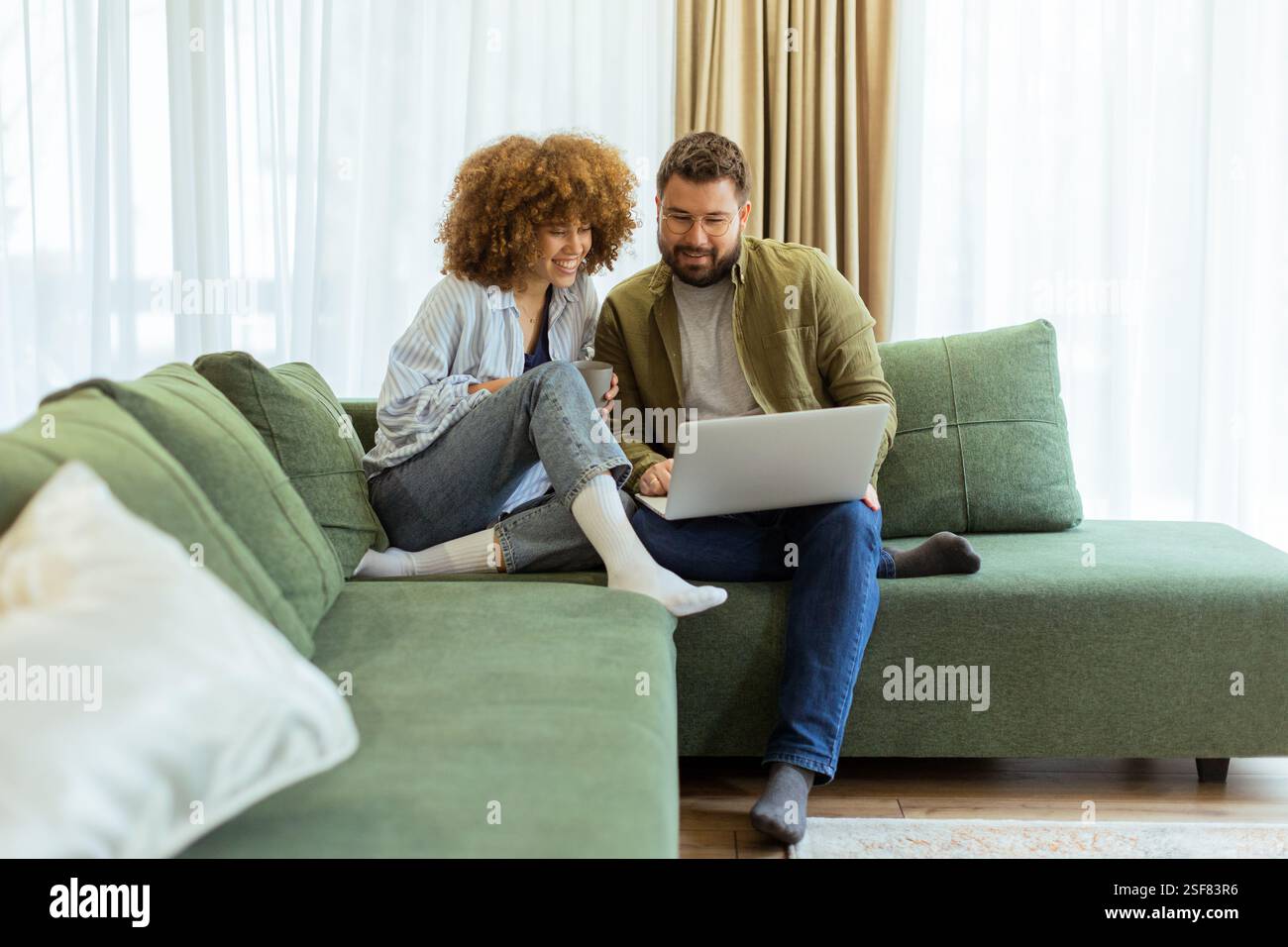 Ein fröhliches Paar sitzt nebeneinander auf einer weichen grünen Couch und führt ein aufregendes Gespräch, während es unter warmem Su auf einem Laptop Ideen durchstöbert Stockfoto