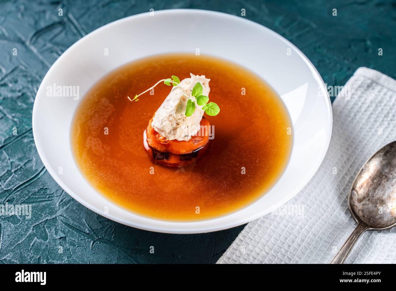 Suppe oder Consomme, dicke Fleischbrühe mit Karotten, Hühnerknödel und Mikrogrün, serviert auf einem weißen Teller auf dunkelgrünem Hintergrund mit Serviette A Stockfoto