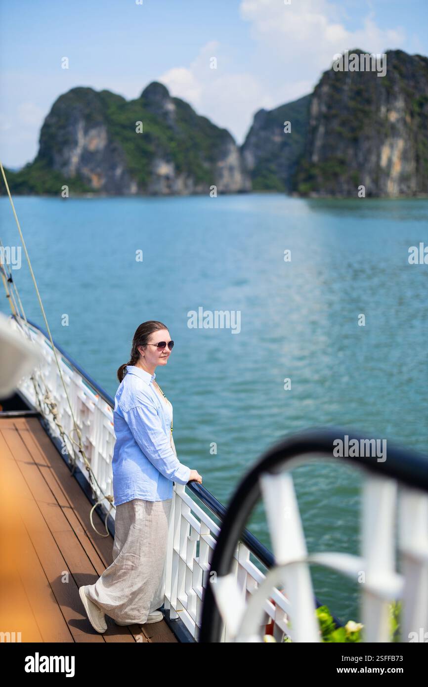Eine Touristenfrau steht auf dem Deck eines Kreuzfahrtschiffes und genießt die malerische Aussicht auf die berühmten Kalksteinklippen der Ha Long Bay und das ruhige blaue Wasser auf einer sonnigen Sonne Stockfoto