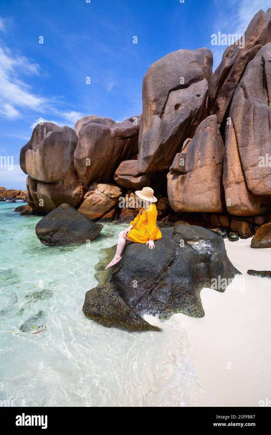 Eine Frau mit breiter Krempe und gelbem Kleid sitzt auf einem großen Felsen am Rand des kristallklaren Wassers Stockfoto