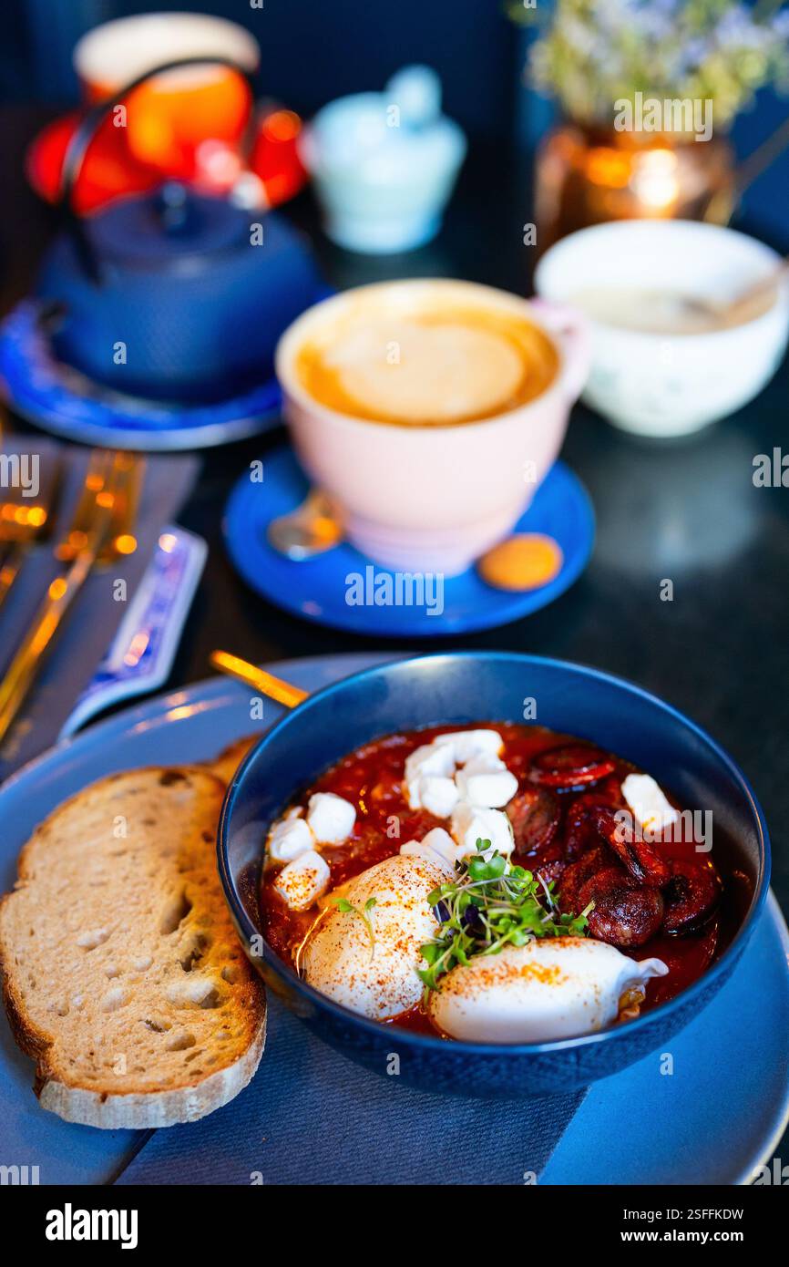 Eine Schüssel Shakshuka mit Chorizo-Scheiben, pochierten Eiern und zerbröckeltem Käse, serviert mit einer Scheibe geröstetem Sauerteigbrot und einer Tasse C Stockfoto