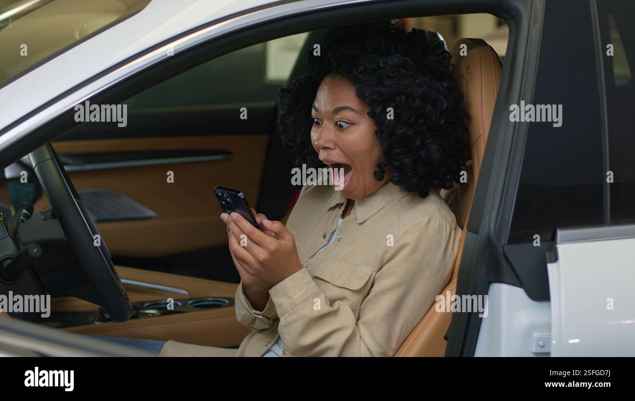 Afroamerikanerin weibliche Fahrerin glückliches Mädchen, das im Auto sitzt und Auto im Autosalon kauft und auf den Bildschirm des Mobiltelefons schaut, erreicht gute Nachrichten Stockfoto