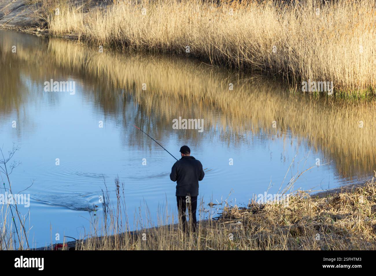 Im Winter fischt eine Person ruhig am Ufer, umgeben von hohen Gräsern und einer friedlichen Wasserspiegelung, und genießt die Ruhe der Natur Stockfoto