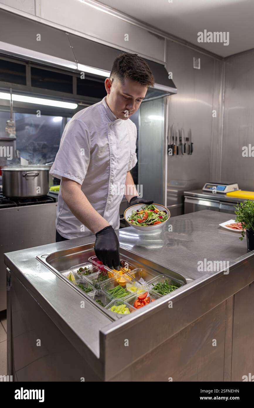 Küchenchef mit Zange und Halteplatte mit frischem Salat über Behältern mit Zutaten in der Restaurantküche Stockfoto