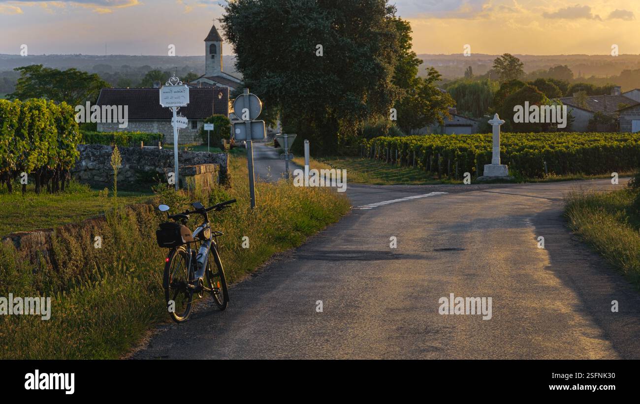 Horizontaler Blick auf die Landschaft entlang einer Landstraße, mit einem Fahrrad auf der linken Seite, einer weißen Ecke des christlichen Kreuzes, einem Kirchturm und Weinbergen Stockfoto