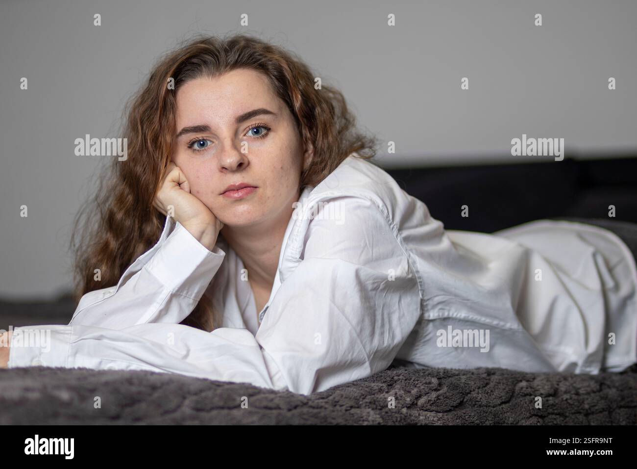 Die junge Frau liegt auf einem Bett, trägt ein weißes Hemd, den Kopf auf der Hand, mit einem kontemplativen Ausdruck Stockfoto