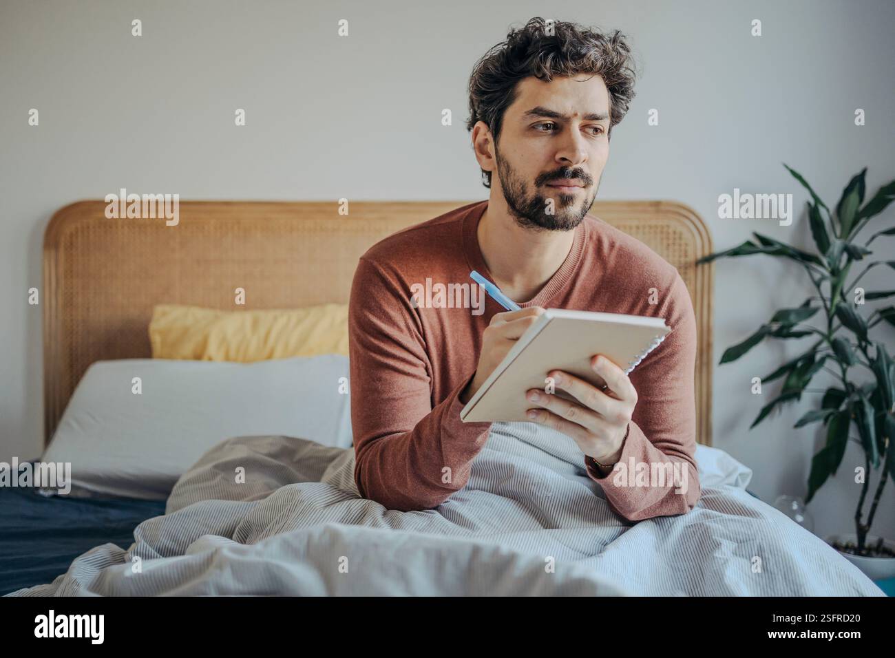 Junger, gutaussehender Mann aus dem Nahen Osten, der im Bett sitzt und in seinem Notizbuch schreibt. Kreativität und Reflexion am Morgen. Stockfoto