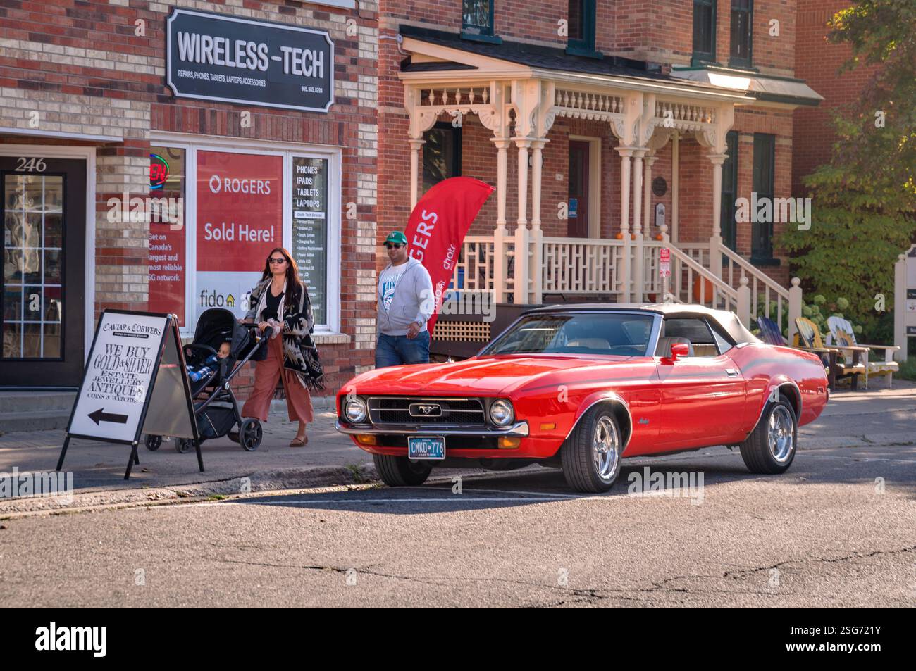 Port Perry, Ontario, Kanada - 2024 10 05: Eine junge Familie, die neben dem roten Ford Mustang Cabrio aus dem Jahr 1971 vor einem alten zweistöckigen Backsteinhaus spaziert Stockfoto