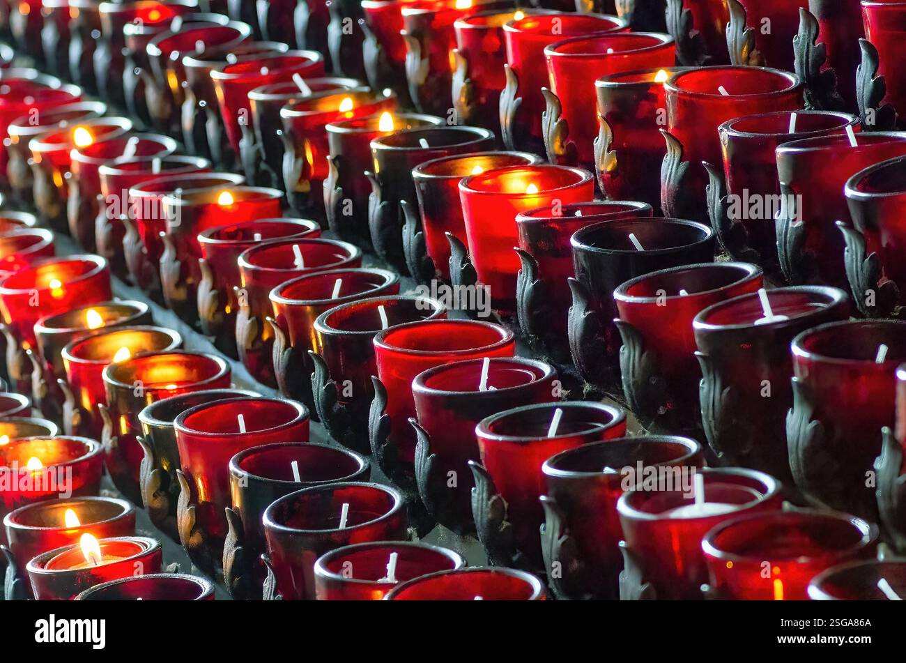 Reihen roter Votivkerzen, einige flackern vor Licht, in einer schwach beleuchteten Umgebung. Die Wiederholung schafft ein warmes und spirituelles Ambiente. Stockfoto