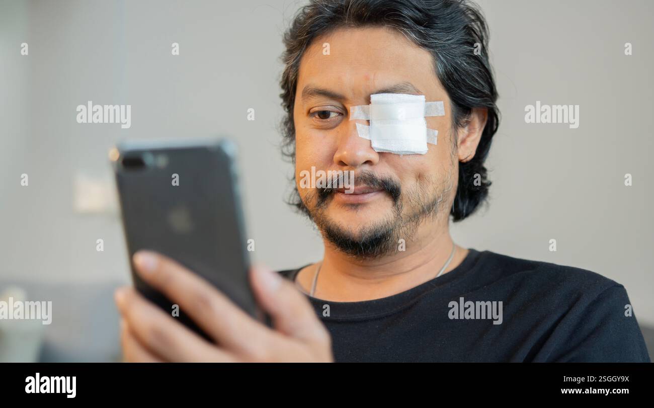 Erwachsener asiatischer Mann mit Augenschmerzen, bedeckt von einem Verband, der auf dem Sofa sitzt und sein Smartphone benutzt. Selektiver Fokus. Stockfoto