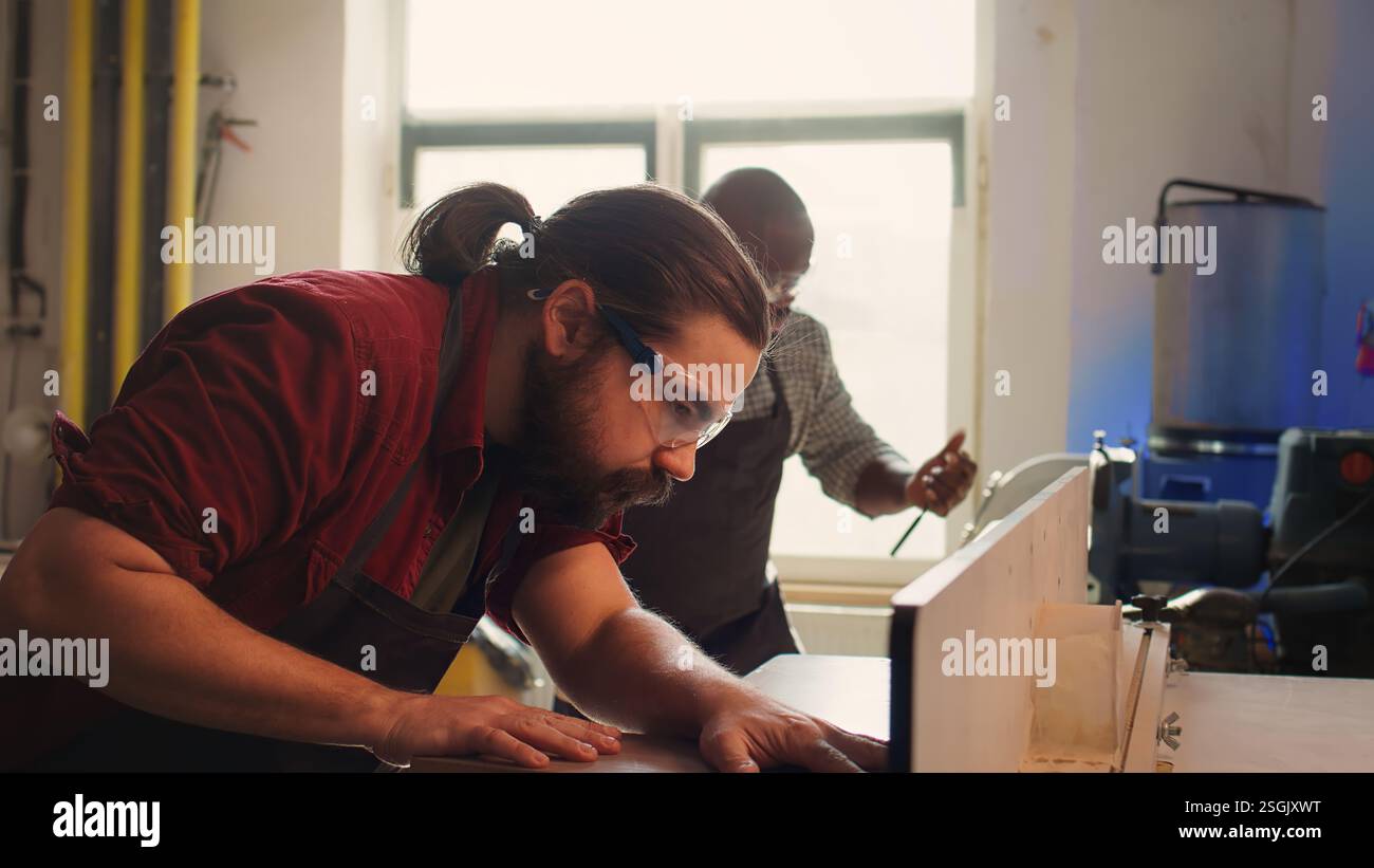 Holzbearbeitungsexperte trägt Schutzkleidung und setzt die Planke in der Spindelmaschine neben dem Kollegen ein. Der CNC-Maschinenbauer verwendet eine Schutzbrille beim Schneiden von Holz mit einem Holzformer, der von seinem Kollegen, Kamera B, unterstützt wird Stockfoto