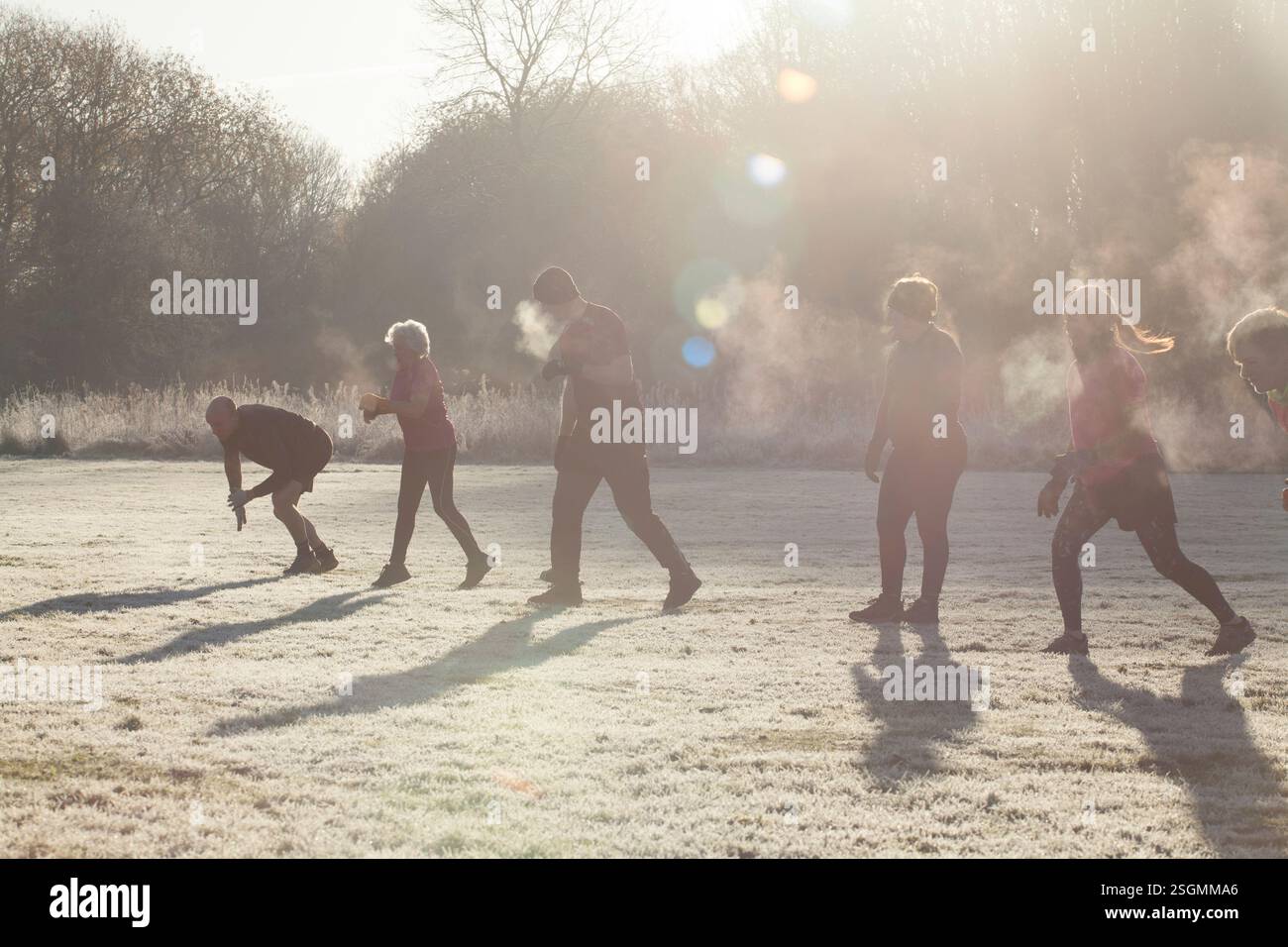 Eine Gruppe von Personen, die sich im Freien auf einem frostigen Feld sportlich betätigen, wobei Sonnenlicht Lichtreflexe und Nebel erzeugt, der aus dem Atem sichtbar ist. Sale Water Park, Manchester, Großbritannien Stockfoto