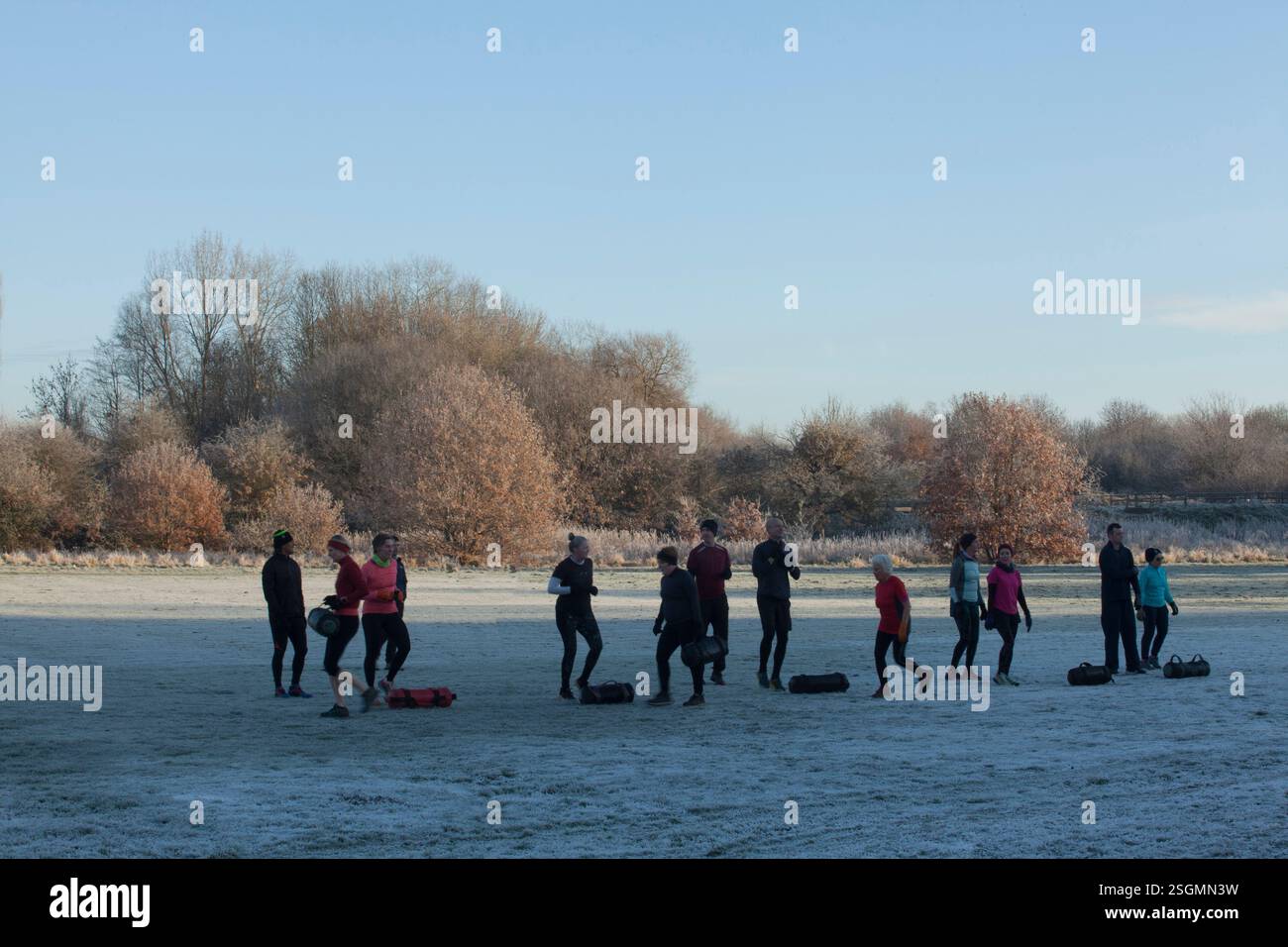 Eine Gruppe von Menschen, die Sportkleidung tragen, übt auf einem frostigen Feld mit verstreuten Bäumen und klarem Himmel Outdoor-Übungen aus. Sale Water Park, Manchester, Großbritannien Stockfoto