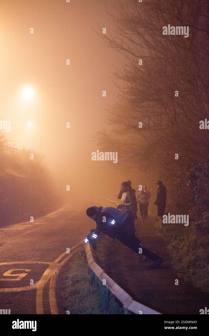 Die Läufer Strecken sich am Abend auf einer nebeligen, schwach beleuchteten Straße. Straßenlaternen strahlen ein warmes Leuchten aus, Sale Water Park, Manchester, Großbritannien Stockfoto