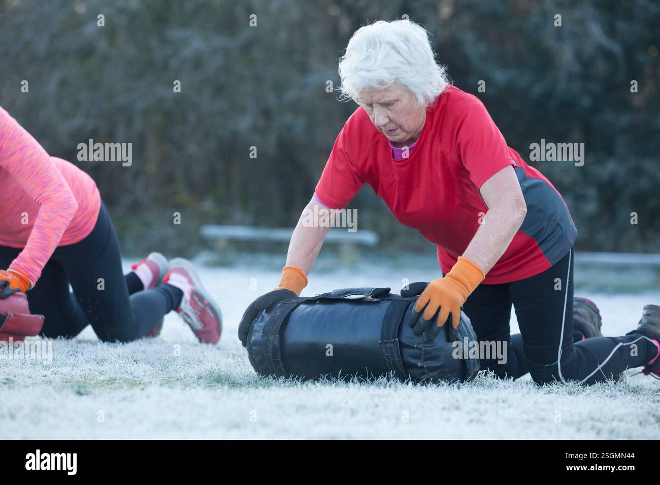 Eine ältere Frau mit weißem Haar, die ein rotes Hemd und orangefarbene Handschuhe trägt, kniet auf frostbedecktem Gras und rollt draußen einen großen schwarzen Übungssandsack. Sale Water Park, Manchester, Großbritannien Stockfoto