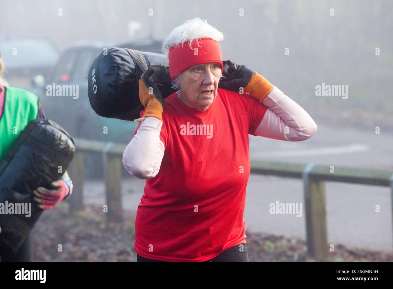 Eine ältere Frau mit weißem Haar, ein rotes Hemd und Stirnband trägt draußen eine schwere schwarze Tasche über den Schultern. Sale Water Park, Manchester, Großbritannien Stockfoto