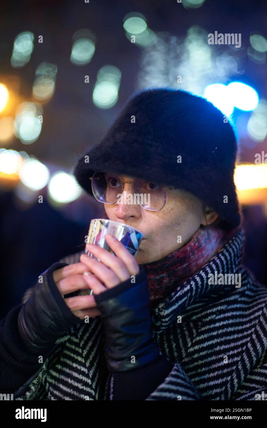 Frau in gestreiftem Mantel und Hut trinkt etwas heißes aus einer Tasse in festlicher, nächtlicher Umgebung. Stockfoto