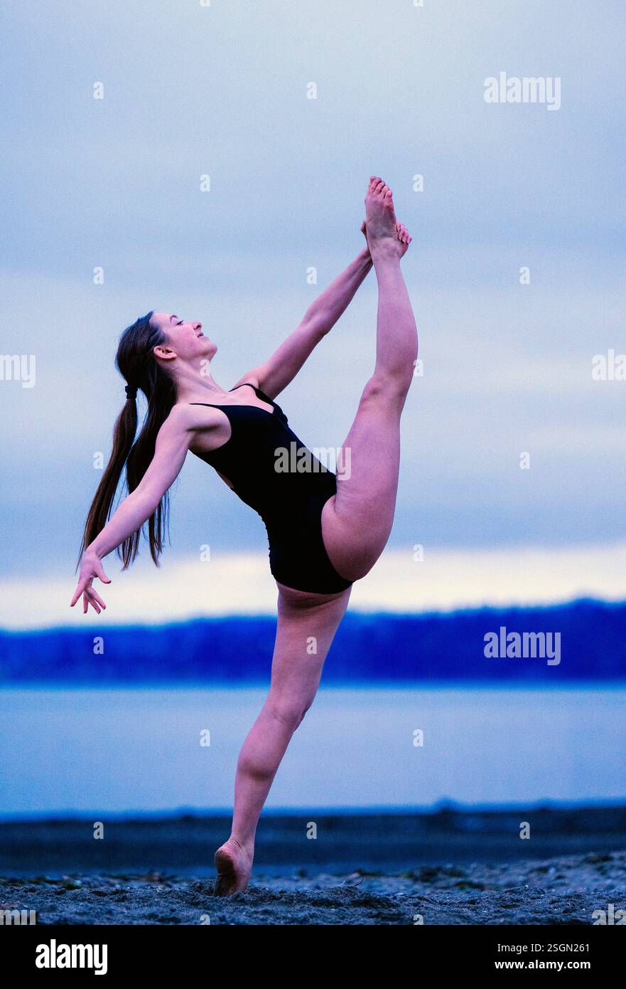 Eine Frau im schwarzen Trikot spielt eine elegante Tanzposition an einem Sandstrand in der Abenddämmerung, USA Stockfoto