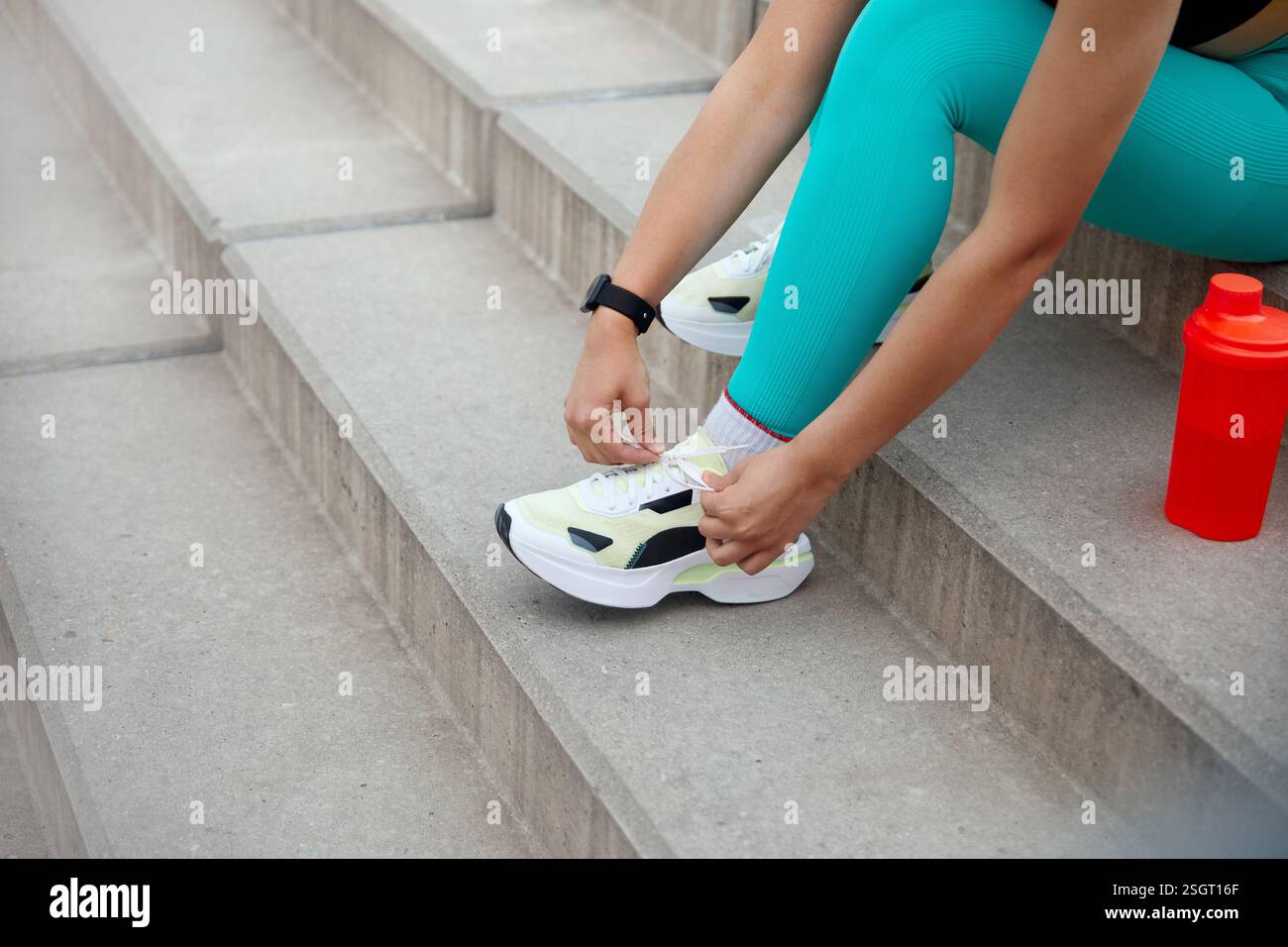 Im Freien sitzt eine Person in lebendiger Trainingskleidung auf Betonstufen und passt ihre Schnürsenkel an, bevor sie eine Trainingseinheit beginnt Stockfoto