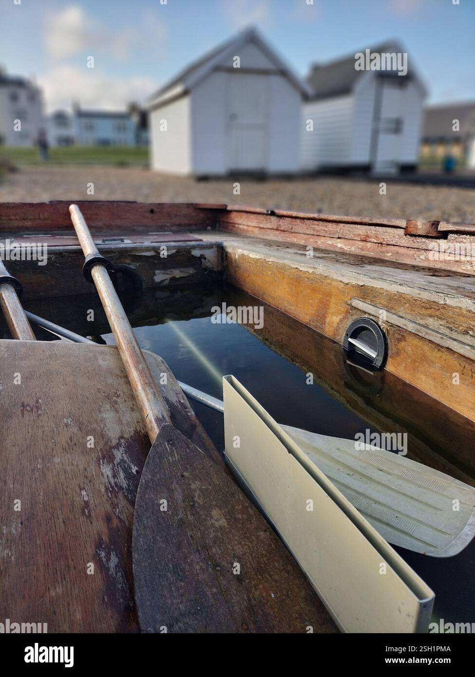 Ein verwittertes Holzboot, das teilweise mit Wasser gefüllt ist und am Ufer mit weißen Strandhütten im Hintergrund ruht. - Smartphone-aufgenommenes Stockfoto