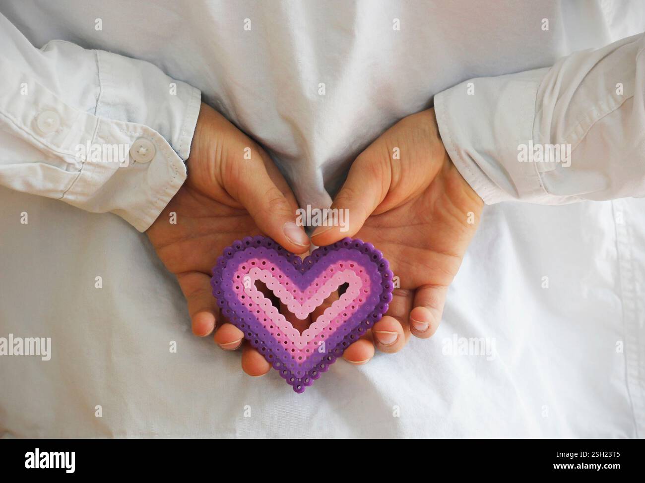 Die Hände des kaukasischen Jungen halten ein rosa lila Plastikherz aus Bügelperlen hinter seinem Rücken. Valentinstag, Muttertag und Vatertag Stockfoto