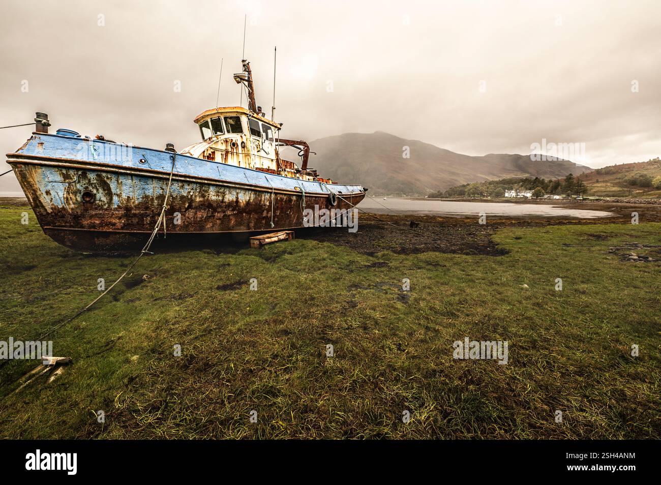Rostiges, verlassenes Fischerboot an der Muddy Shore mit Bergen und bewölktem Himmel Stockfoto