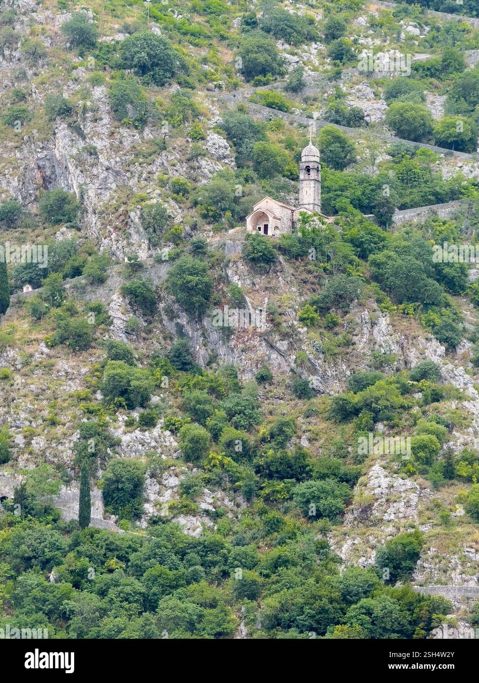 Kotor, Montenegro - 2. Juli 2024: Kirche unserer Lieben Frau von Remedy auf dem hohen grauen Berghang über der historischen Altstadt. Flecken von grünen Büschen Stockfoto