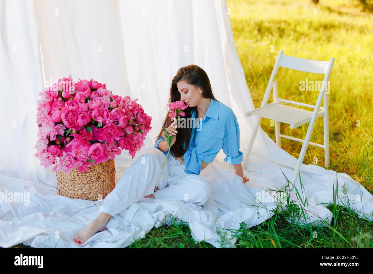 Eine stilvolle Frau in einem blauen Hemd und einer weißen Hose sitzt draußen barfuß auf einem weißen Stoff und riecht nach rosa Pfingstrose. Ein großer Korb mit Blumen und ein Weißer Stockfoto