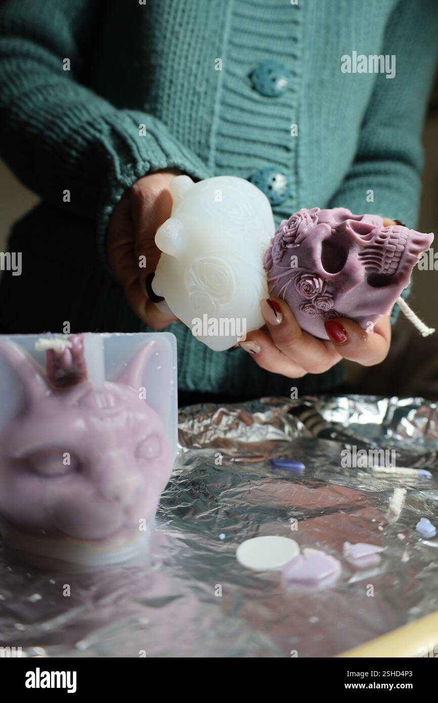 Eine junge Frau, die eine Schädelkerze auflöst, neben einer fertigen lila Kerzenform in Form einer Katze. DIY-Kerzen, Wachsbasteln Stockfoto