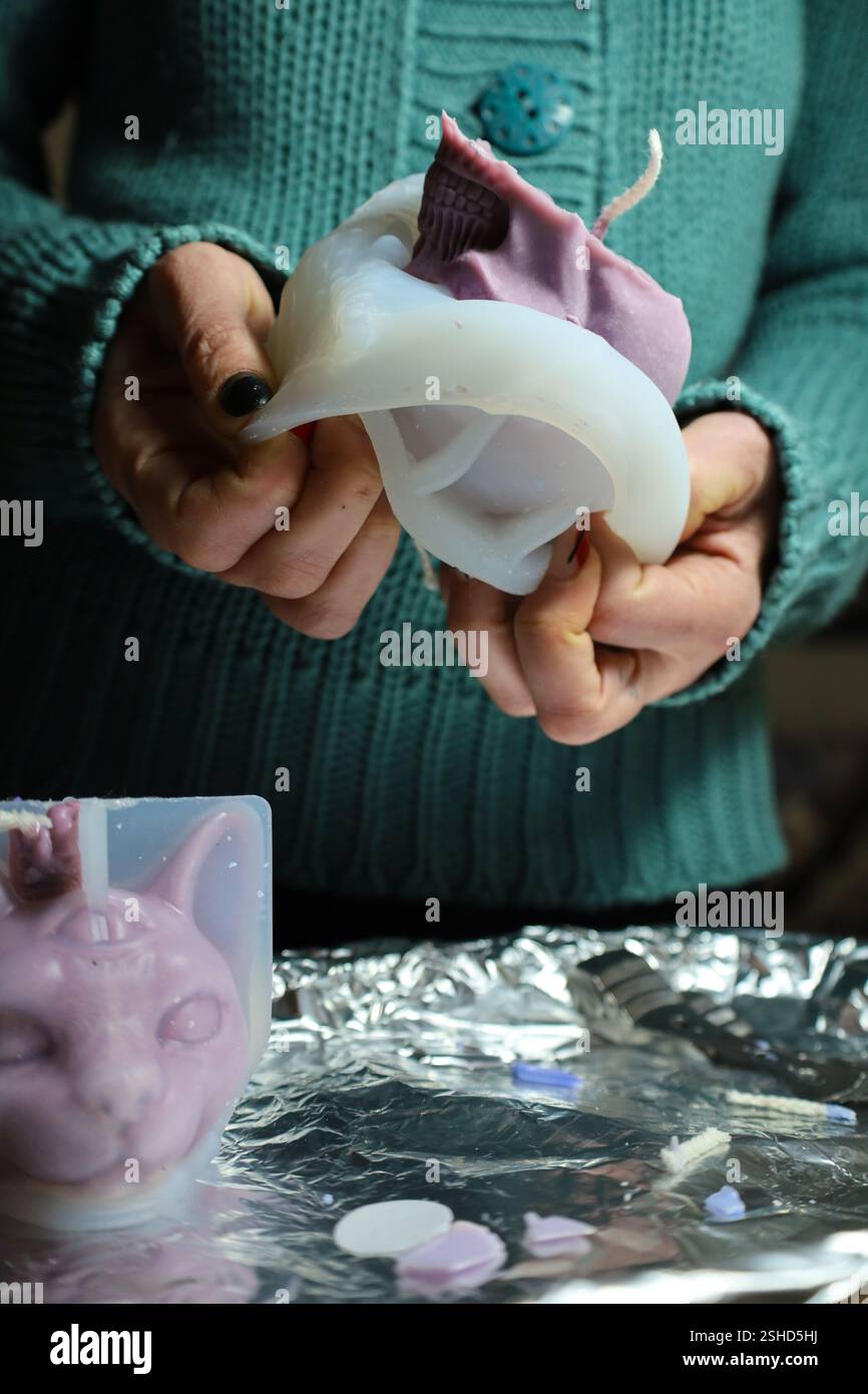 Eine junge Frau, die eine weiße Silikonform für eine Kerze entformt, neben einer fertigen lila Kerzenform in Form einer Katze. DIY-Kerzen, Wachsbasteln Stockfoto