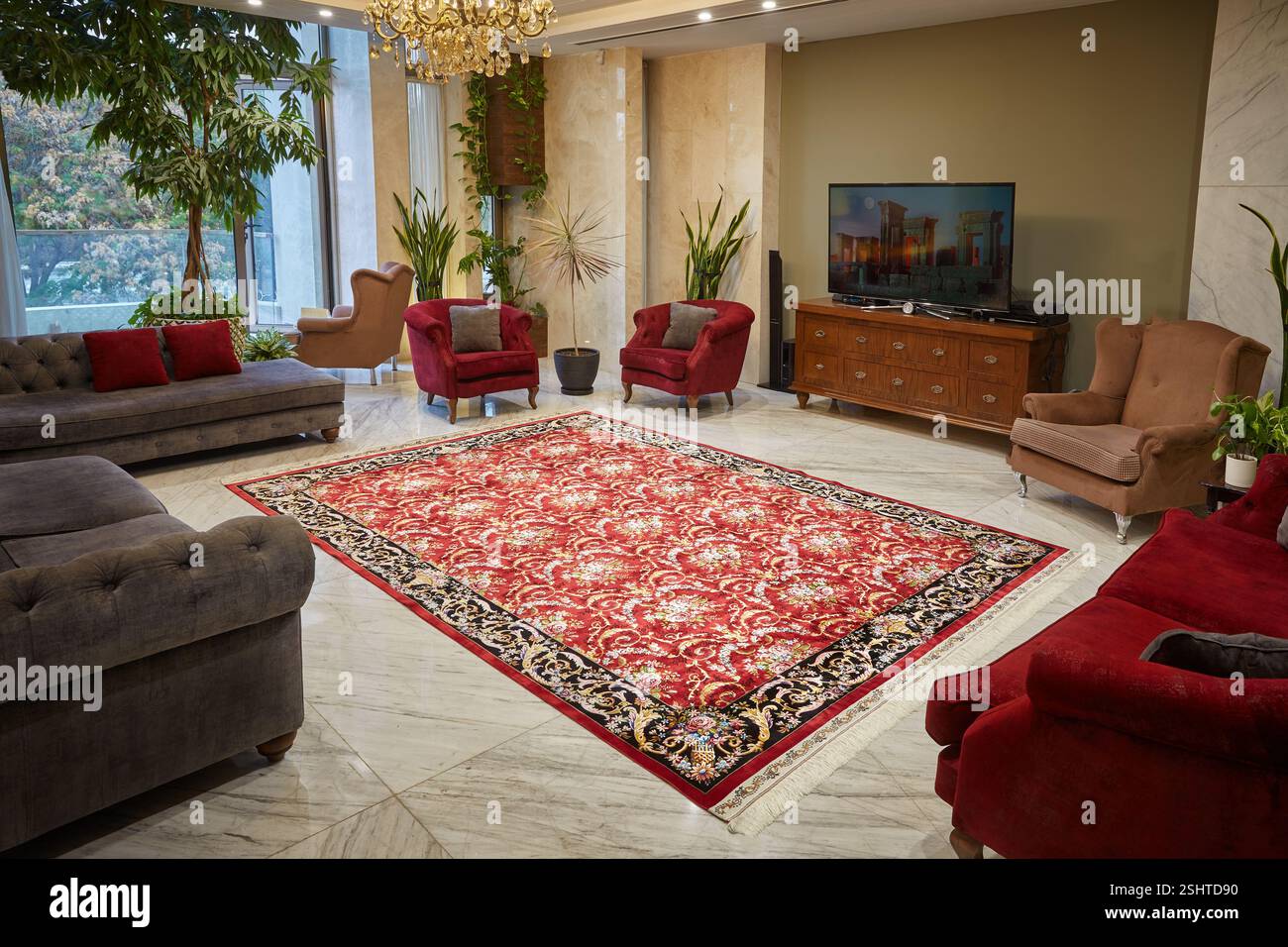 Modernes Wohnzimmer mit einem roten persischen Teppich mit einem komplizierten Medaillon, Marmorböden und üppigen Innenpflanzen. Stockfoto