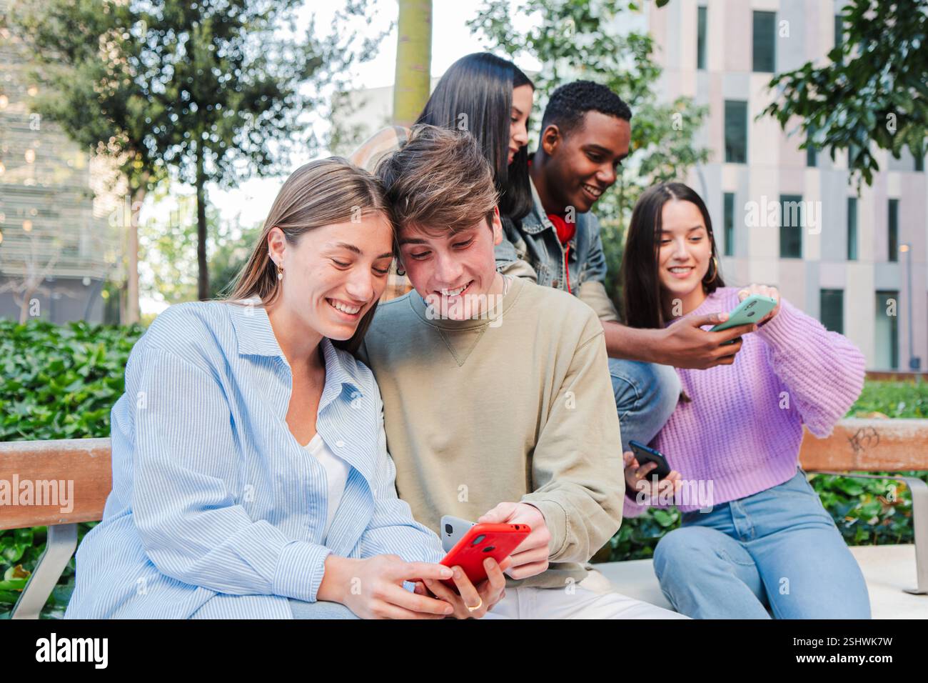 Fröhliche Gruppe junger Freunde, die mit ihren Handys interagieren und spannende Momente und Lachen teilen. Teenager konzentrierten sich auf ihr Mobiltelefon, genossen Erlebnisse und schufen gemeinsam Verbindungen. Hochwertige Fotos Stockfoto