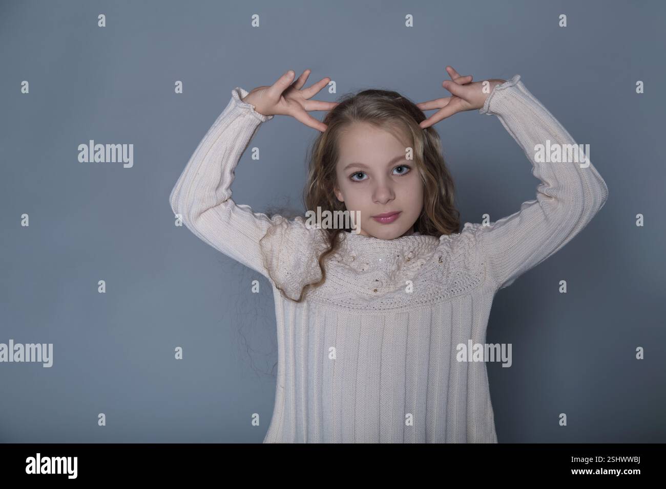 Ein Mädchen mit langen Haaren hebt spielerisch die Hände in einer selbstbewussten Pose und trägt einen stylischen Pullover. Dieses Foto zeigt sowohl ihre lustige Persönlichkeit als auch die Stockfoto