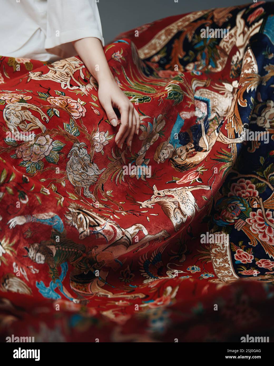 Roter Teppich mit Tiermotiven und Blumenmustern hängt elegant ab, gehalten von einer Frau in einem weißen Kleid Stockfoto