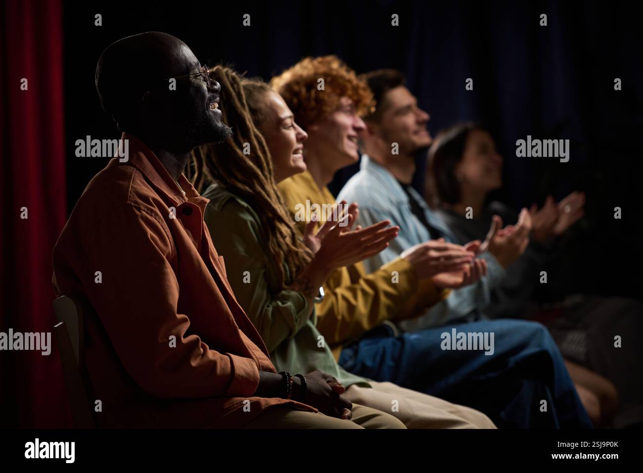Gruppe von verschiedenen Personen applaudiert einer Vorstellung mit fröhlichem Ausdruck, während sie in einer schwach beleuchteten Umgebung sitzen, die Zweisamkeit und Spaß hervorhebt Stockfoto