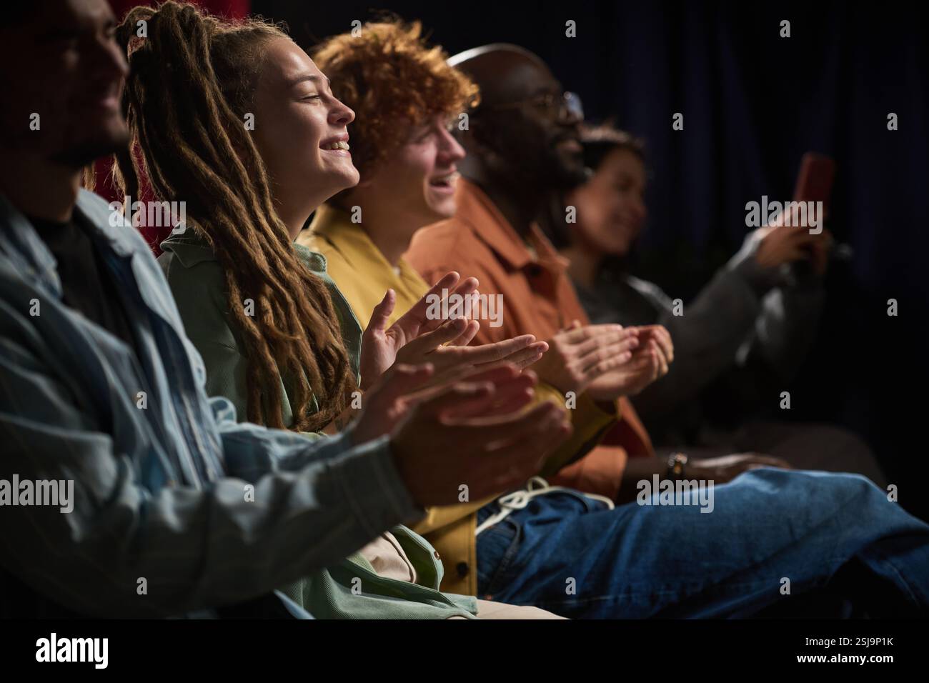 Eine Gruppe verschiedener Personen verschiedener Ethnien genießt und applaudiert eine Vorstellung, sitzt eng zusammen in einem verdunkelten Raum und teilt gemeinsame Erfahrungen und Verbindungen Stockfoto