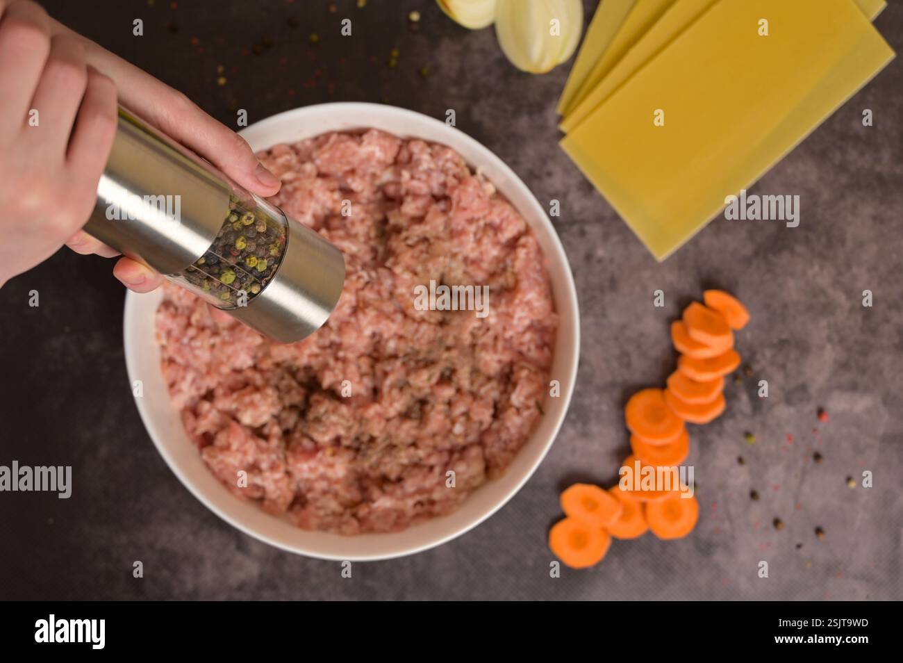 Blick von oben rohes Hackfleisch in weißer Schüssel, Frauenhände halten Pfeffermühle. Lasagne Nudelblätter, Zwiebeln. Karotte, Pfeffer. Schwarzer Hintergrund Stockfoto